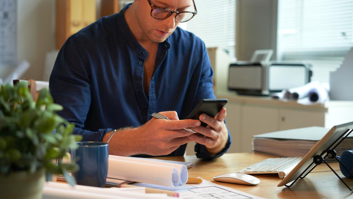 caucasian man sitting desk with rolled up papers plans using smartphone